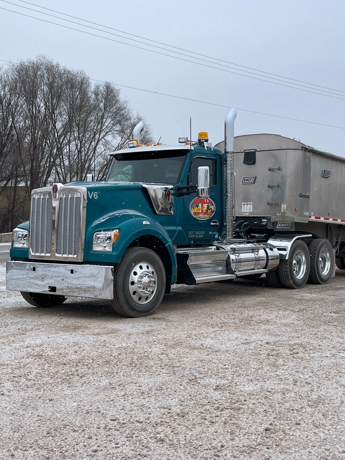 Teal Kenworth semi with an aluminum East Genesis end dump trailer parked on a gravel lot in light snow
